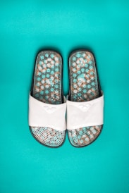 A pair of white slide sandals featuring a textured sole with a mosaic pattern in shades of turquoise, brown, and white, set against a vibrant turquoise background.
