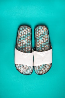 A pair of white slide sandals featuring a textured sole with a mosaic pattern in shades of turquoise, brown, and white, set against a vibrant turquoise background.