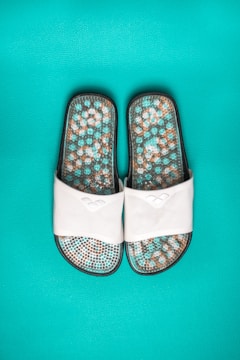 A pair of white slide sandals featuring a textured sole with a mosaic pattern in shades of turquoise, brown, and white, set against a vibrant turquoise background.