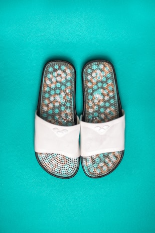 A pair of white slide sandals featuring a textured sole with a mosaic pattern in shades of turquoise, brown, and white, set against a vibrant turquoise background.