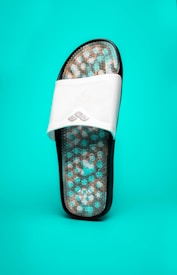 A single sandal with a white strap and a textured sole featuring a pattern of small rounded bumps in blue and beige. The sandal is positioned upright against a bright teal background.
