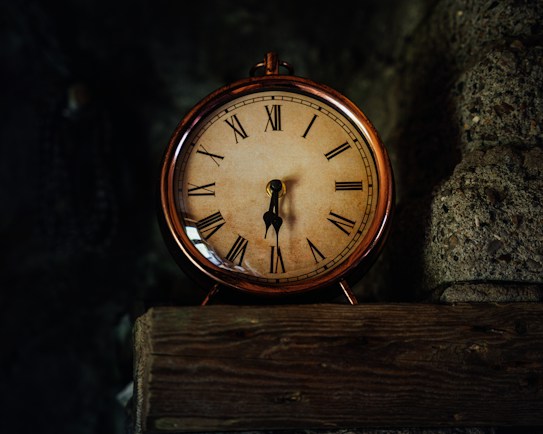 A vintage clock with a bronze frame sits on a rustic wooden shelf. The clock face features Roman numerals and classic black clock hands, set against a textured stone wall in dim lighting.