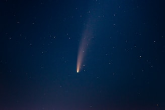 A comet with a bright, glowing core and a long, trailing tail traverses a star-filled night sky. The tail creates a faint, slightly curved line of light extending from the core, set against a dark blue backdrop.