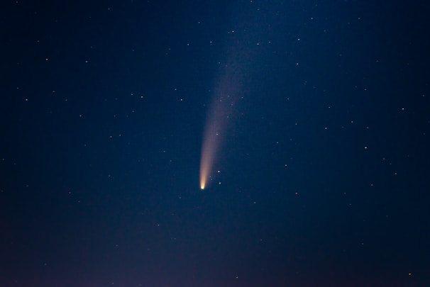 A comet with a bright, glowing core and a long, trailing tail traverses a star-filled night sky. The tail creates a faint, slightly curved line of light extending from the core, set against a dark blue backdrop.