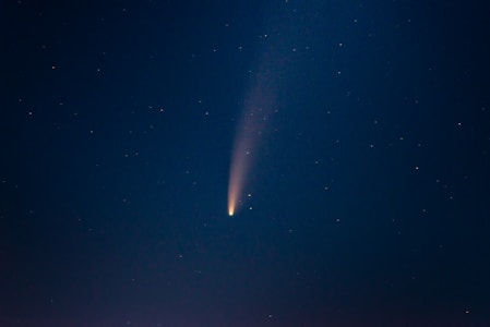 A comet with a bright, glowing core and a long, trailing tail traverses a star-filled night sky. The tail creates a faint, slightly curved line of light extending from the core, set against a dark blue backdrop.