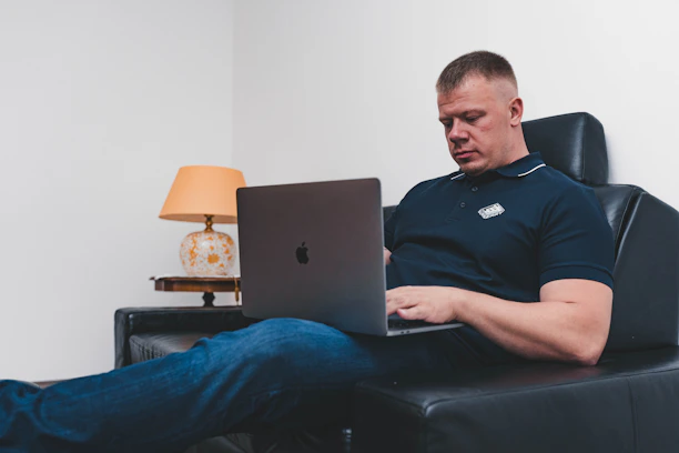 man in black polo shirt sitting on black leather couch using macbook