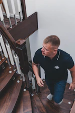 man in black crew neck t-shirt and blue denim jeans standing beside brown wooden staircase