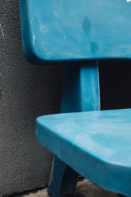 A close-up view of a blue bench positioned against a textured dark wall. The bench has a slightly worn surface with visible scratches, and the angle captures the corner and a portion of the seat, showing its sturdy base.