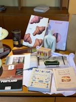 The image features a collection of books and magazines displayed on a wooden table, including titles about food, lifestyle, and cities. Items such as a ceramic vase with a fern design, a box of tea, and a tin labeled with hot chocolate are also present. A fabric, seemingly a cloth or napkin, lies beneath these objects.