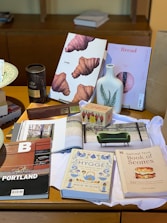 The image features a collection of books and magazines displayed on a wooden table, including titles about food, lifestyle, and cities. Items such as a ceramic vase with a fern design, a box of tea, and a tin labeled with hot chocolate are also present. A fabric, seemingly a cloth or napkin, lies beneath these objects.