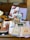 The image features a collection of books and magazines displayed on a wooden table, including titles about food, lifestyle, and cities. Items such as a ceramic vase with a fern design, a box of tea, and a tin labeled with hot chocolate are also present. A fabric, seemingly a cloth or napkin, lies beneath these objects.