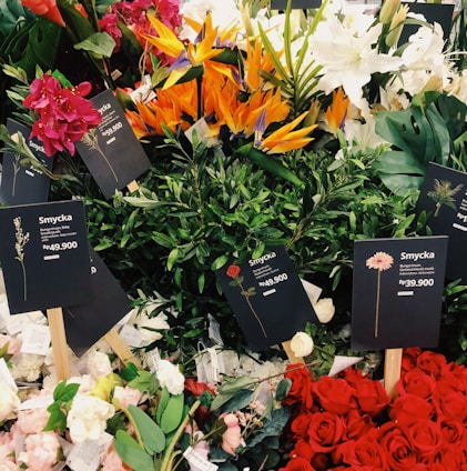 A variety of colorful flower bouquets arranged together prominently. Several types of flowers including red roses, white lilies, orange Birds of Paradise, and pink and red flowers are visible. Black price tags with the label 'Smycka' and prices in Rp currency are placed among the flowers.