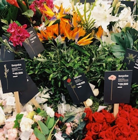 A variety of colorful flower bouquets arranged together prominently. Several types of flowers including red roses, white lilies, orange Birds of Paradise, and pink and red flowers are visible. Black price tags with the label 'Smycka' and prices in Rp currency are placed among the flowers.
