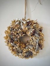 A decorative wreath made of dried leaves and flowers is hanging on a wall. It features a rustic ribbon tied at the top, adding to its natural aesthetic.
