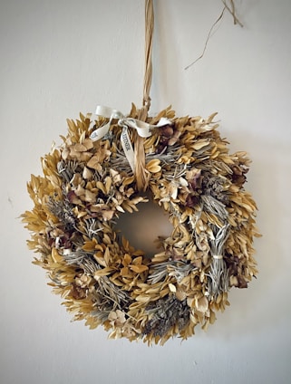 A decorative wreath made of dried leaves and flowers is hanging on a wall. It features a rustic ribbon tied at the top, adding to its natural aesthetic.
