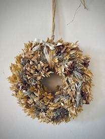 A decorative wreath made of dried leaves and flowers is hanging on a wall. It features a rustic ribbon tied at the top, adding to its natural aesthetic.