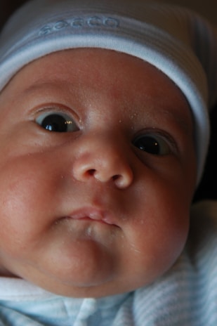 Close-up of a baby's face with chubby cheeks and wide eyes. The baby is wearing a light blue beanie and a matching onesie. The soft lighting casts gentle shadows across the baby's features, adding warmth to the image.