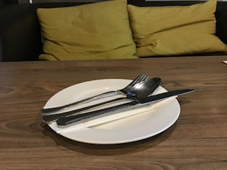 A wooden table is set with a white plate, placed centrally, holding silver cutlery including a knife, spoon, and fork. Behind the table are plush yellow-green cushions on a dark seating area, creating a cozy and inviting atmosphere.