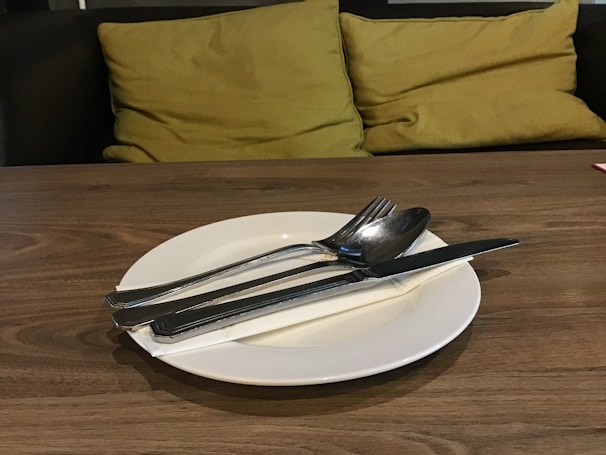 A wooden table is set with a white plate, placed centrally, holding silver cutlery including a knife, spoon, and fork. Behind the table are plush yellow-green cushions on a dark seating area, creating a cozy and inviting atmosphere.