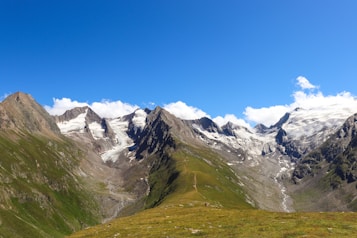 Snow-capped mountains with rugged peaks surround a lush green valley. A narrow trail leads through the landscape, and a bright blue sky with scattered clouds adds to the serene atmosphere.