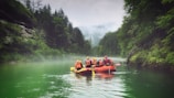 A group of people wearing helmets and life jackets are rafting in green water. They are in a red inflatable raft surrounded by lush, green forested cliffs. Mist hangs over the calm river, creating a serene and adventurous atmosphere.