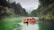 A group of people wearing helmets and life jackets are rafting in green water. They are in a red inflatable raft surrounded by lush, green forested cliffs. Mist hangs over the calm river, creating a serene and adventurous atmosphere.