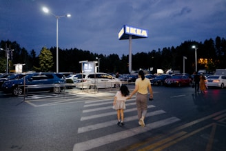 A mother and child are crossing a parking lot towards an IKEA store in the evening. The sky is dark, and the parking lot is almost full with parked cars and a few people walking. Street lamps and the IKEA sign are illuminating the scene.