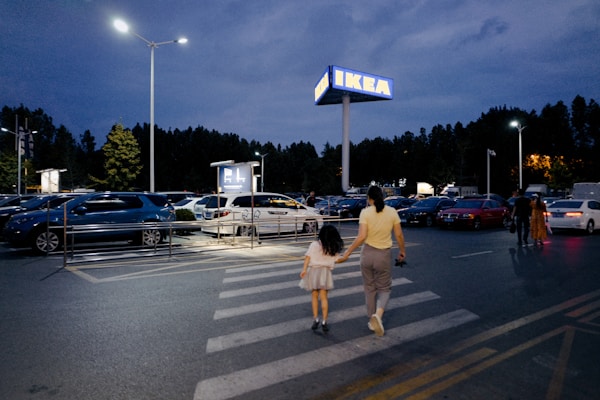 A mother and child are crossing a parking lot towards an IKEA store in the evening. The sky is dark, and the parking lot is almost full with parked cars and a few people walking. Street lamps and the IKEA sign are illuminating the scene.