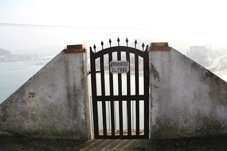 A wrought iron gate set in a concrete wall overlooks a river or body of water. The gate has a sign that reads 'PROHIBIDO EL PASO', indicating no entry. In the background, there's a misty view of a bridge, buildings, and a road.