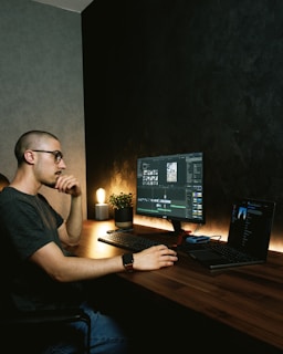 A person is seated at a wooden desk working on a computer with a large monitor displaying what appears to be video editing software. There is a laptop beside the monitor, a small potted plant, a lamp, and various tech accessories. The background is a dark textured wall, and the lighting gives a warm, focused atmosphere.