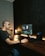 A person is seated at a wooden desk working on a computer with a large monitor displaying what appears to be video editing software. There is a laptop beside the monitor, a small potted plant, a lamp, and various tech accessories. The background is a dark textured wall, and the lighting gives a warm, focused atmosphere.