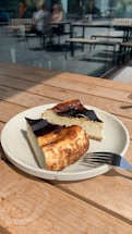 A ceramic plate holds two slices of cheesecake with a dark, caramelized top. The plate is on a wooden table, with sunlight casting shadows. In the background, there are blurred images of people seated at tables outside.