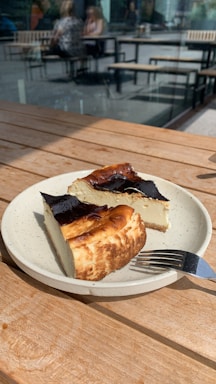 A ceramic plate holds two slices of cheesecake with a dark, caramelized top. The plate is on a wooden table, with sunlight casting shadows. In the background, there are blurred images of people seated at tables outside.