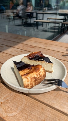 A ceramic plate holds two slices of cheesecake with a dark, caramelized top. The plate is on a wooden table, with sunlight casting shadows. In the background, there are blurred images of people seated at tables outside.