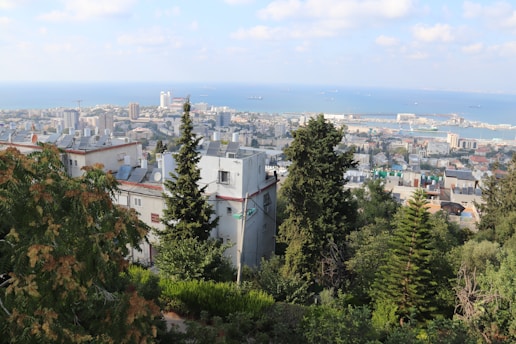 A panoramic view of a coastal cityscape featuring a blend of urban and natural elements. In the foreground, lush green trees and vegetation are visible, with residential buildings partially obscured among the foliage. The middle ground showcases a densely built city with numerous buildings, some high-rises, and solar panels on rooftops. The background features a vast expanse of blue sea with ships visible in the water, under a sky scattered with clouds.