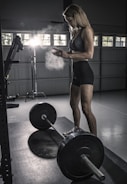 A person in athletic attire stands in a dimly lit gym, preparing to lift a barbell. Chalk dust is visible in the air, illuminated by a bright light in the background. The setting is industrial, with large windows and weightlifting equipment nearby.
