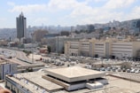 Urban landscape featuring a cluster of modern and commercial buildings, with a prominent high-rise structure in the foreground. A curved road lined with parked vehicles winds through the scene. In the background, a hilly area is covered with densely packed buildings.