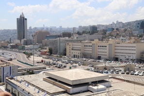 Urban landscape featuring a cluster of modern and commercial buildings, with a prominent high-rise structure in the foreground. A curved road lined with parked vehicles winds through the scene. In the background, a hilly area is covered with densely packed buildings.