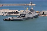 A large naval ship docked at a harbor, surrounded by calm blue water. Buildings and various equipment are visible on the shore, indicating a port area. The ship has a sleek design with multiple antennas and radar equipment on top.
