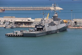 A large naval ship docked at a harbor, surrounded by calm blue water. Buildings and various equipment are visible on the shore, indicating a port area. The ship has a sleek design with multiple antennas and radar equipment on top.