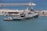 A large naval ship docked at a harbor, surrounded by calm blue water. Buildings and various equipment are visible on the shore, indicating a port area. The ship has a sleek design with multiple antennas and radar equipment on top.