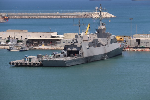 A large naval ship docked at a harbor, surrounded by calm blue water. Buildings and various equipment are visible on the shore, indicating a port area. The ship has a sleek design with multiple antennas and radar equipment on top.