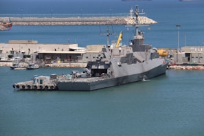 A large naval ship docked at a harbor, surrounded by calm blue water. Buildings and various equipment are visible on the shore, indicating a port area. The ship has a sleek design with multiple antennas and radar equipment on top.