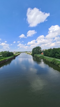 A serene river flows between lush green banks, reflecting the bright blue sky dotted with fluffy white clouds. Tall trees line the riverbanks, and a small red-roofed building is visible in the distance, adding a touch of color to the verdant landscape.