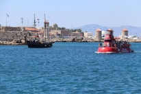 A scenic coastal view featuring a modern tourist vessel and a traditional sailing ship on a clear blue sea. In the background, there is a stone fortress with flags and several windmills, set against a backdrop of distant mountains. The sunlight creates a vibrant and lively atmosphere.