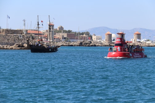 A scenic coastal view featuring a modern tourist vessel and a traditional sailing ship on a clear blue sea. In the background, there is a stone fortress with flags and several windmills, set against a backdrop of distant mountains. The sunlight creates a vibrant and lively atmosphere.