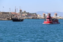 A scenic coastal view featuring a modern tourist vessel and a traditional sailing ship on a clear blue sea. In the background, there is a stone fortress with flags and several windmills, set against a backdrop of distant mountains. The sunlight creates a vibrant and lively atmosphere.