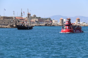 A scenic coastal view featuring a modern tourist vessel and a traditional sailing ship on a clear blue sea. In the background, there is a stone fortress with flags and several windmills, set against a backdrop of distant mountains. The sunlight creates a vibrant and lively atmosphere.