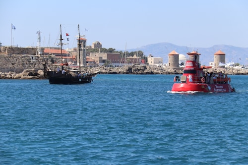 A scenic coastal view featuring a modern tourist vessel and a traditional sailing ship on a clear blue sea. In the background, there is a stone fortress with flags and several windmills, set against a backdrop of distant mountains. The sunlight creates a vibrant and lively atmosphere.