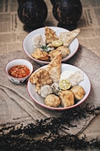 Two bowls filled with various types of fried and steamed snacks, garnished with a slice of lime and served alongside a small dish of red chili sauce. The setup is on a rustic cloth, with an underlying surface of newspaper print and decorative elements in the background.